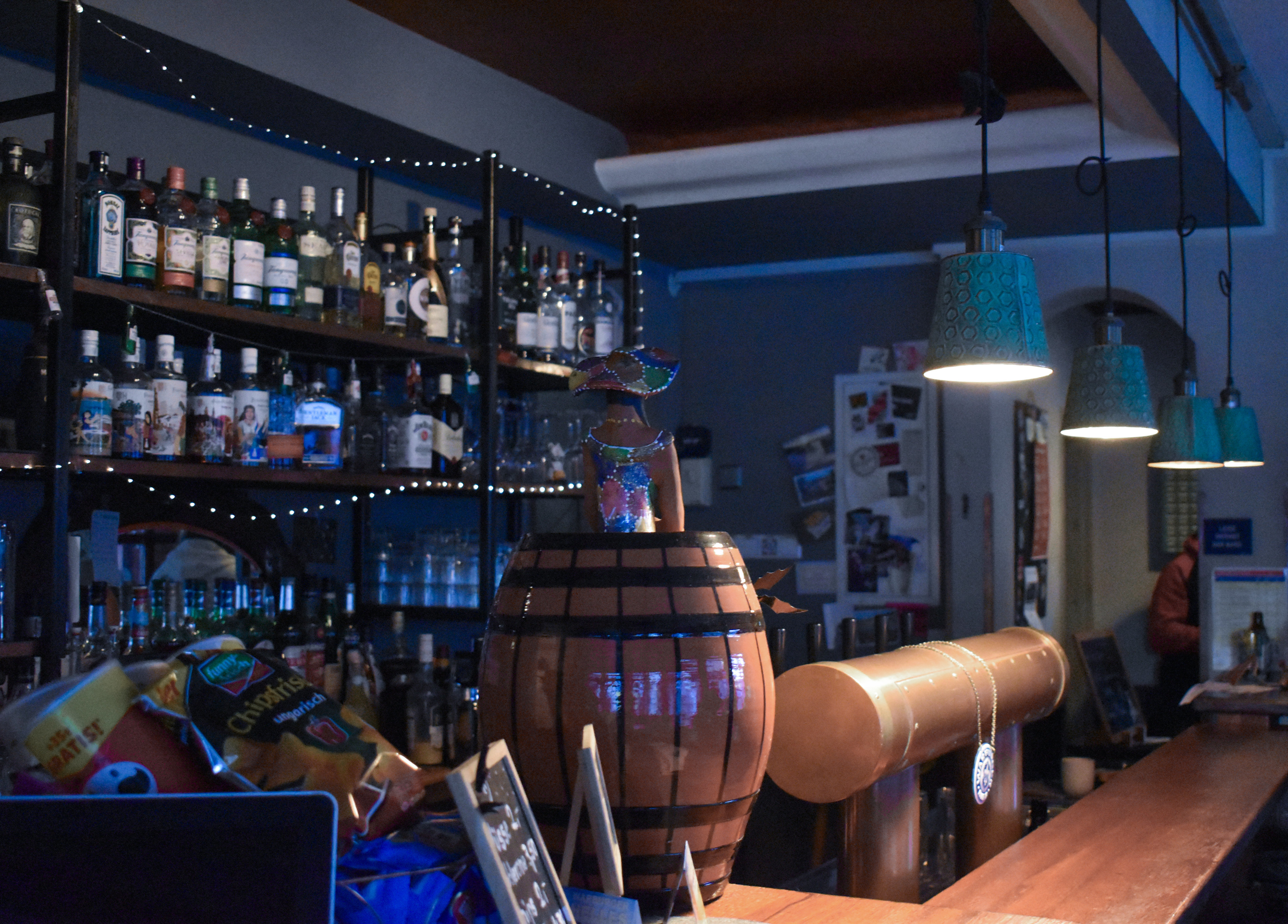 Bild von der Bar der Kneipe AKKA, hinter der Bar stehen Regale mit vielen Flaschen und leuchtenden Lichterketten, im Vordergrund ein Korb mit Snacks und ein hölzernes Fass mit einer dekorativen Figur
