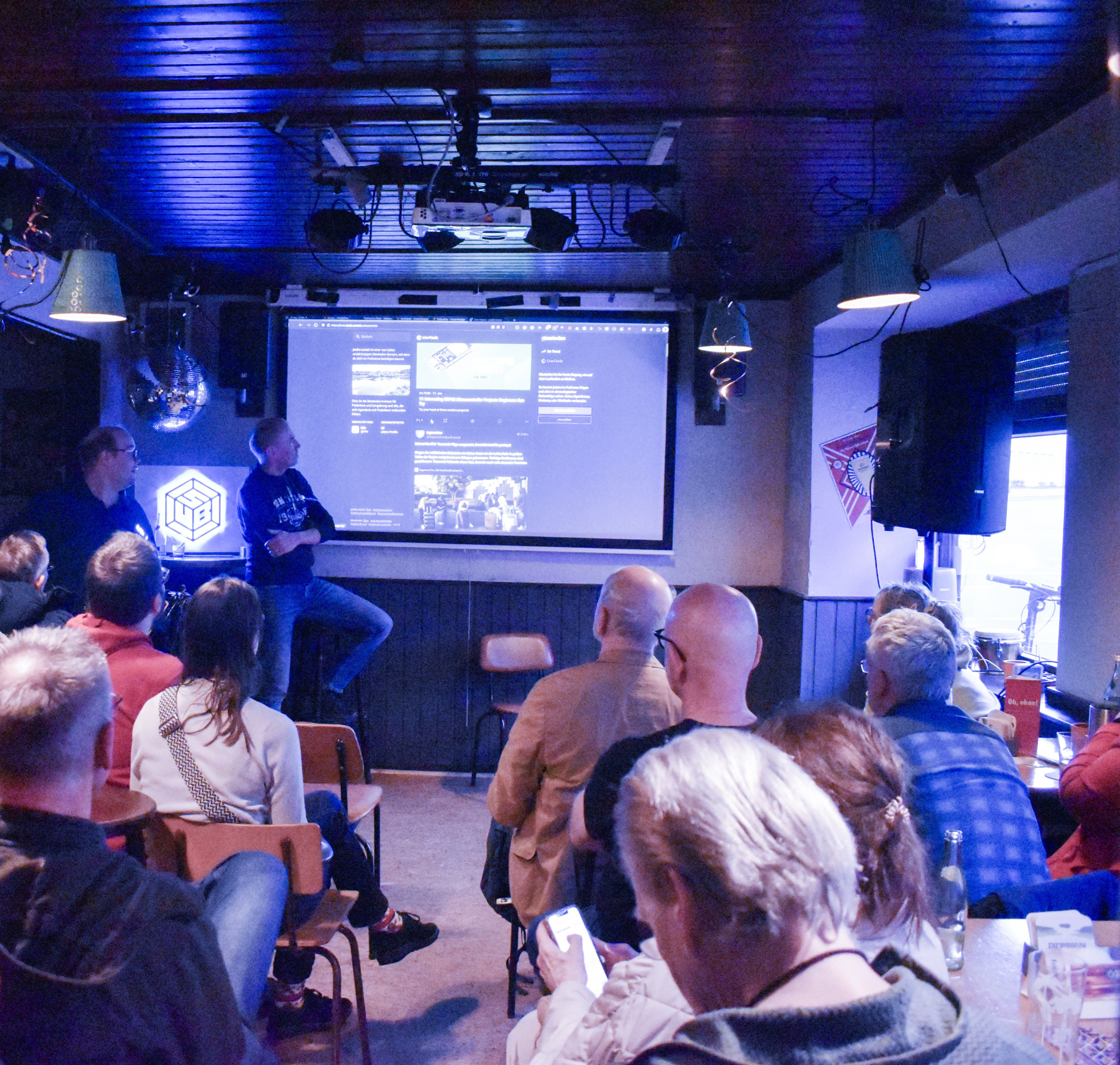 Bild von der Veranstaltung zum 2. DI-Day in Paderborn im AKKA, zwei Redner stehen vor einer Leinwand und präsentieren eine Mastodon-Seite, mehrere Personen sitzen vor ihnen und hören dem Vortrag zu.